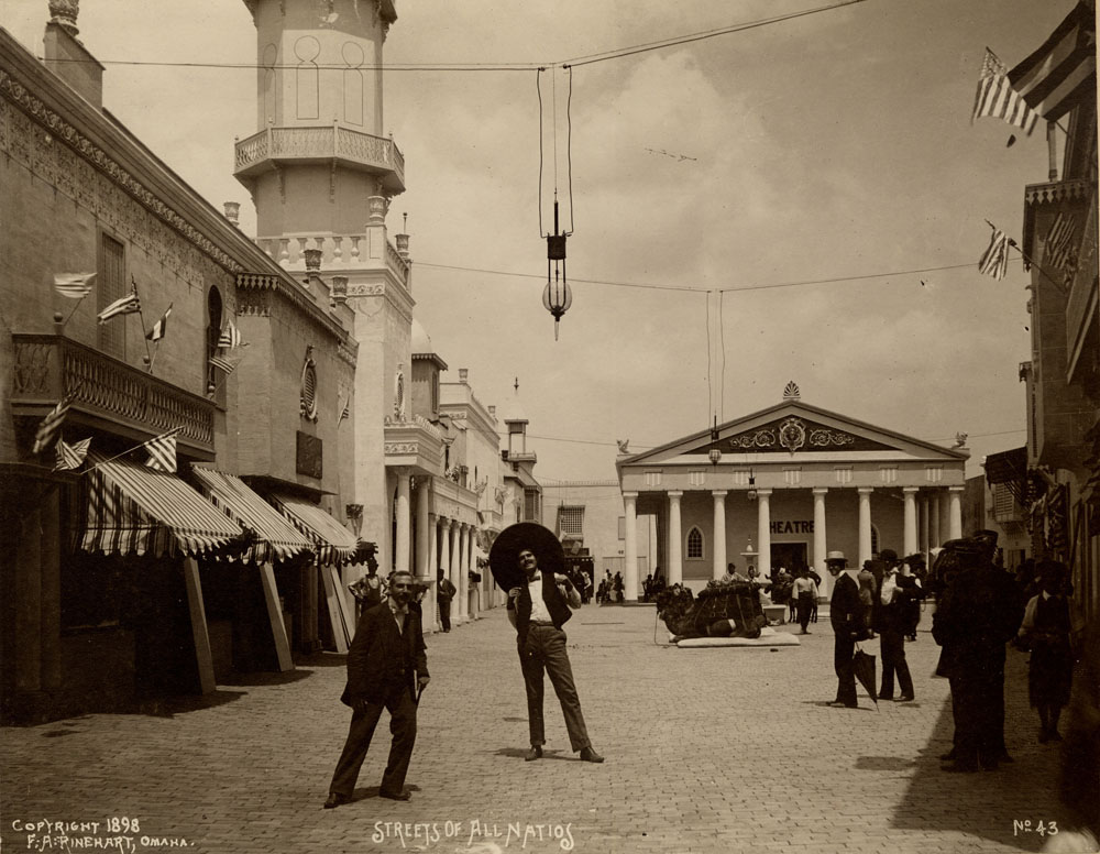 Two men pose at Streets of All Nations. Theater in background. One man wears a sombrero. Light crowd in background.