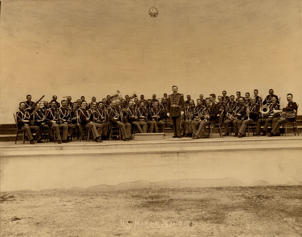 U.S. Marine Band seated on stage of Band Stand, in uniform and with intstruments.