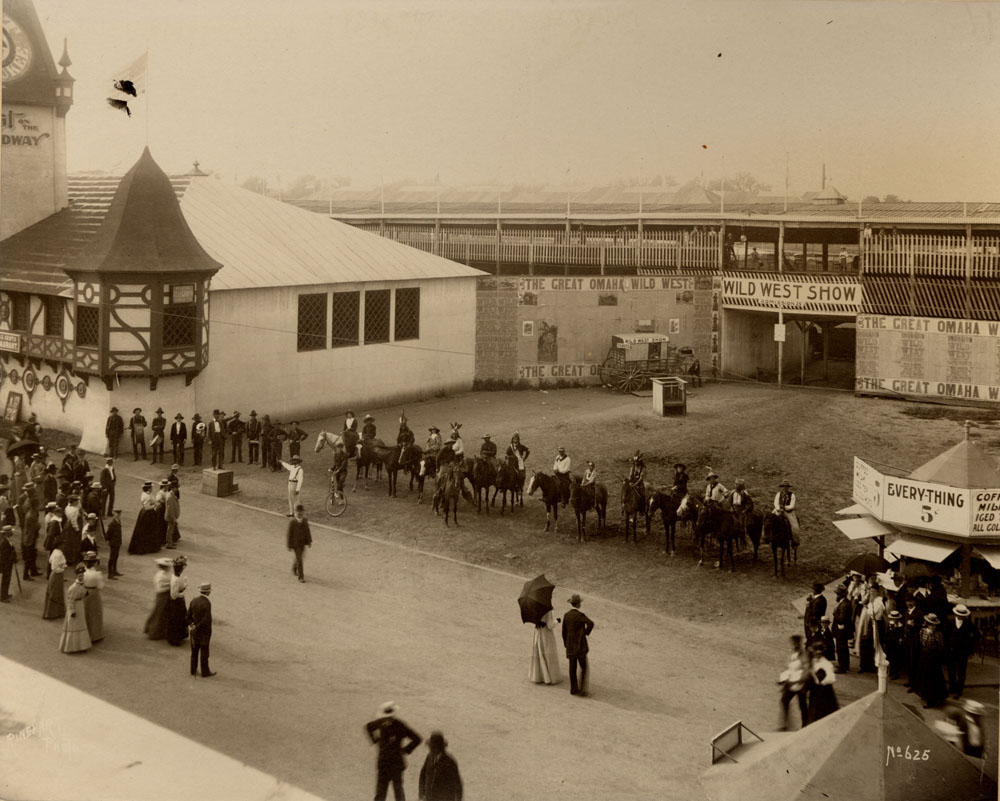 Wild West show entrance. Performers line up on horseback. Pabst Pavilion to the northwest. Light crowd gathered.