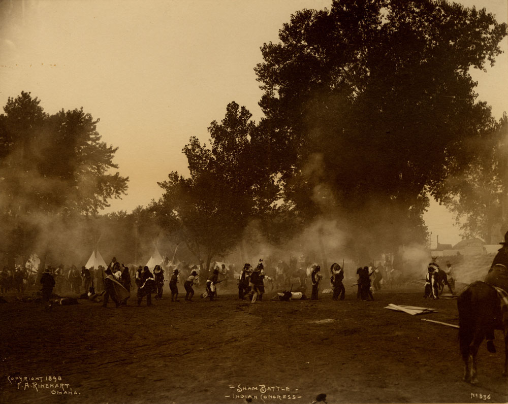 Indian Congress participants engage in mock battle. Gun smoke fills the air. Man on horse in front right foreground. Bleacher seating to the right.
