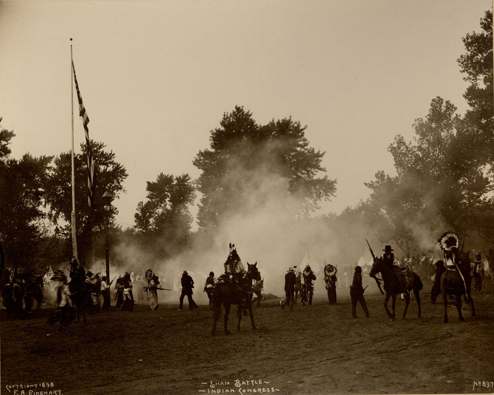 Indian Congress participants engage in mock battle. Gun smoke fills the air. Flag pole with American Flag.
