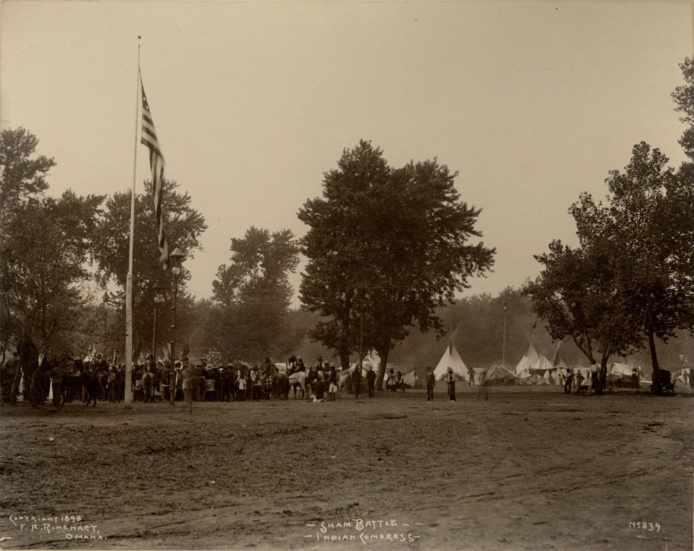 Indian Congress participants gathered for mock battle. Flag pole with American flag to the left. Teepees in background.