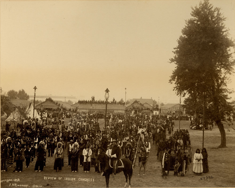 Overview of Indian Congress. Participants gather in foreground. Captain W. A. Mercer on horseback in foreground, spectators crowd in background, standing in front of building and on rooftops.