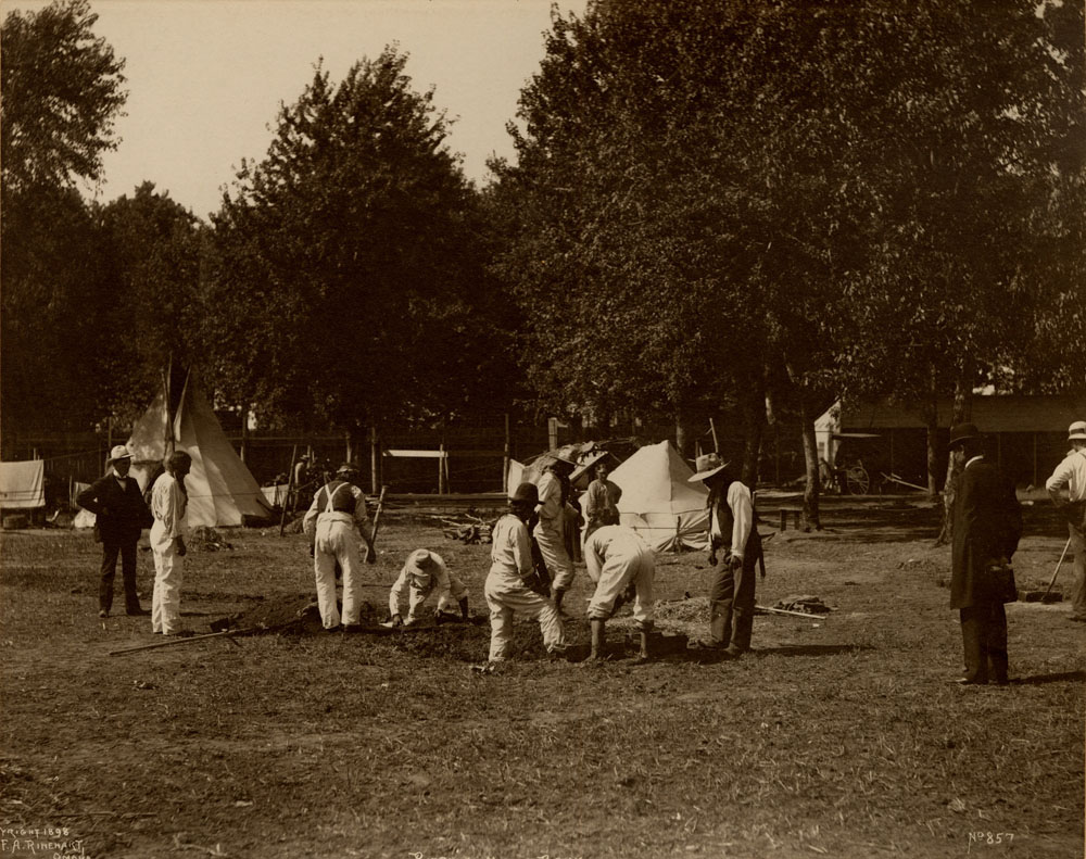Indian Congress particitpants digging at an encampment. Teepees nearby. Carriage in background. A couple spectators observe.