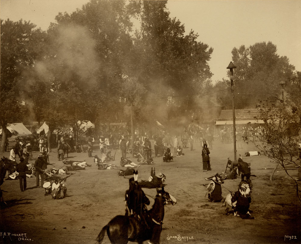 Indian Congress Participants engaged in mock battle. Gun smoke fills the air. Bleachers and tents in the background. Man on horse in foreground.