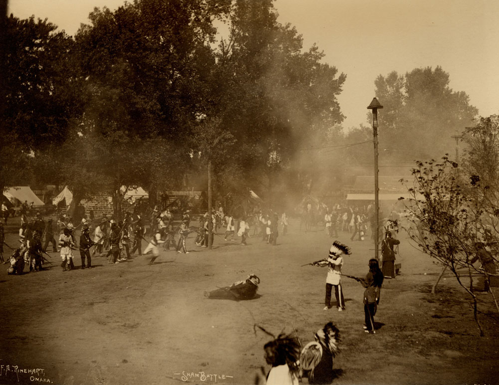 Indian Congress Participants engaged in mock battle. Gun smoke fills the air. Bleachers and tents in the background.