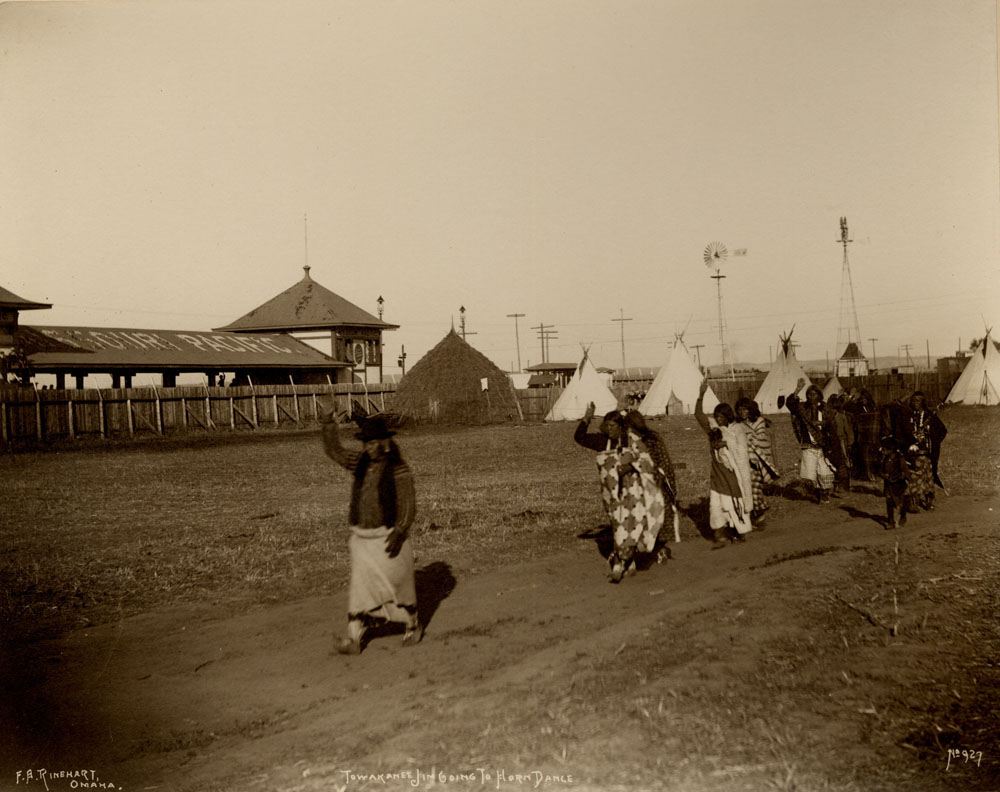 Chief Towohkonie Jim (Wichita) going to Horn Dance, followed by his wife and others. Missouri Pacific Railroad Station, wigwam, teepees, and windmill in background.