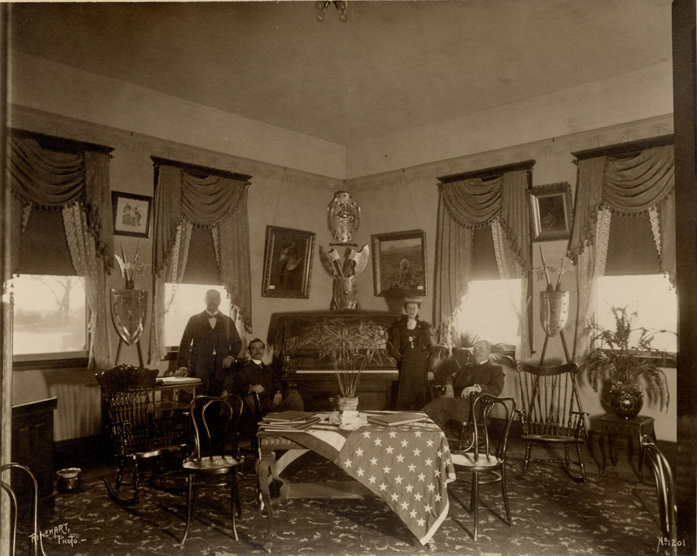 Knights of Pythias display. Sitting room, with chairs, side tables, and potted plants. Three men and a woman pose next to a piano. An American flag is drapped over a coffee table. Framed artwork and Knights of Pythias shields adorn the walls. 