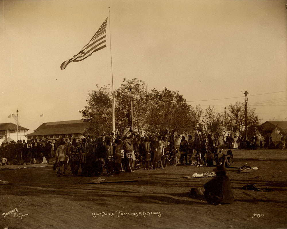 Arapahoe and Cheyennes Delegates at Indian Congress, gathered around a flag pole with American flag, performing Crow Dance. Spectators in background. Man seated in foreground. Transportation and Agricultural Implements Building, Giant See-Saw, Dairy Building, and teepees in background.