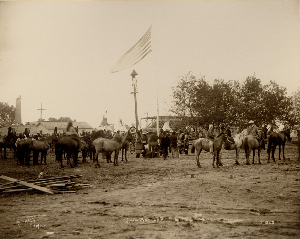 Indian Congress Participants gathered for mock battle, many on horseback. Flag pole with American flag in center. Teepees in background, buildings, and bleachers in background.