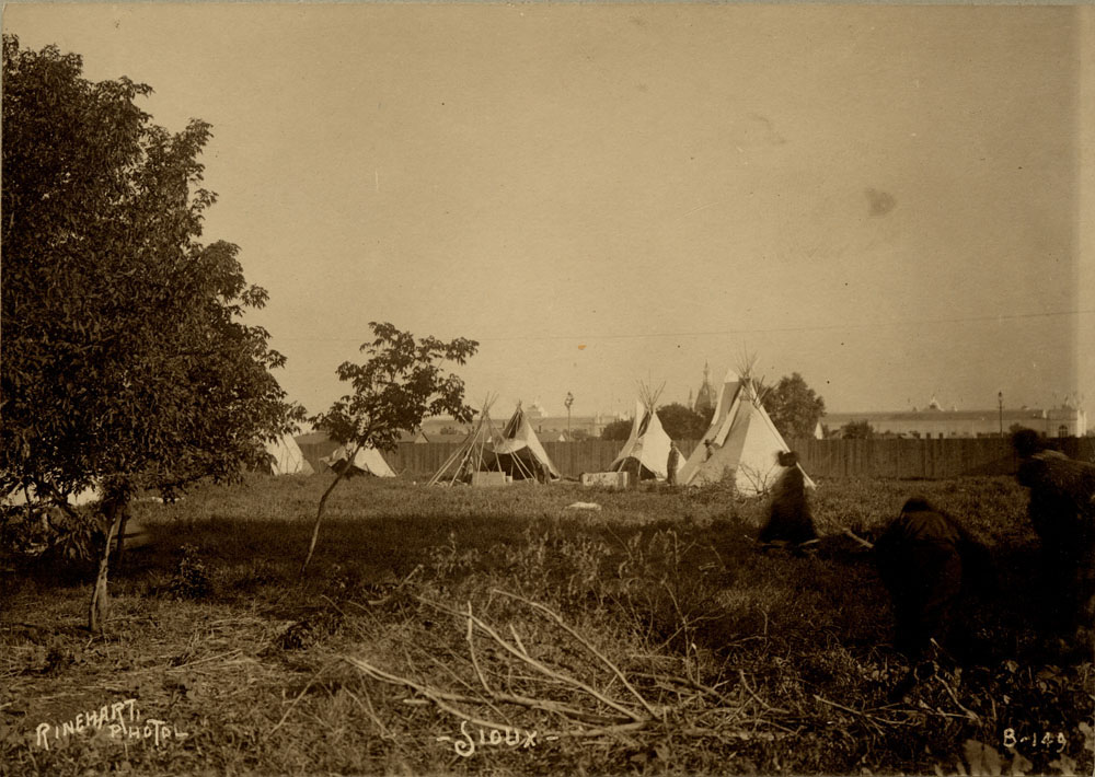 Sioux Encampment at Indian Congress. Trees and branches in foreground. A few teepees in background, and three blurred people in right foreground.