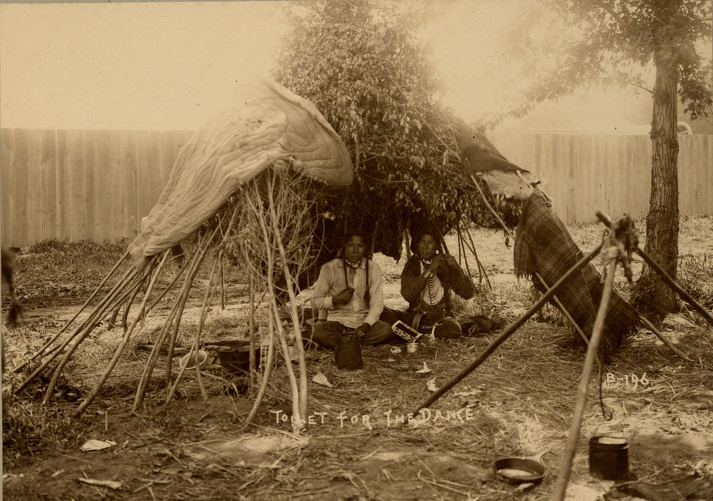 Indian Congress Participants. Two men preparing for dance inside structure made of branches and blankets.