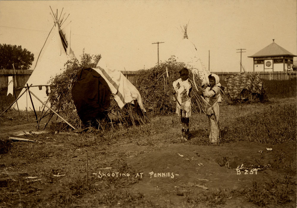 Two young Indian Congress Participants shoot arrows at pennies next to various dwellings at Assiniboine encampment. Missouri Pacific Railroad Station in background.