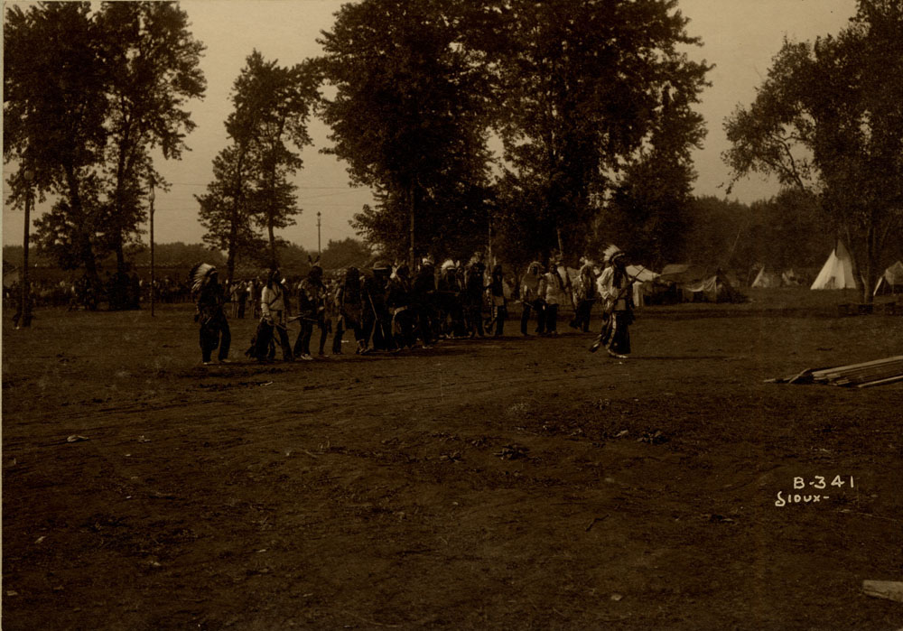 Sioux Delegates of the Indian Congress stand at encampment. Teepees and wigwams in background.
