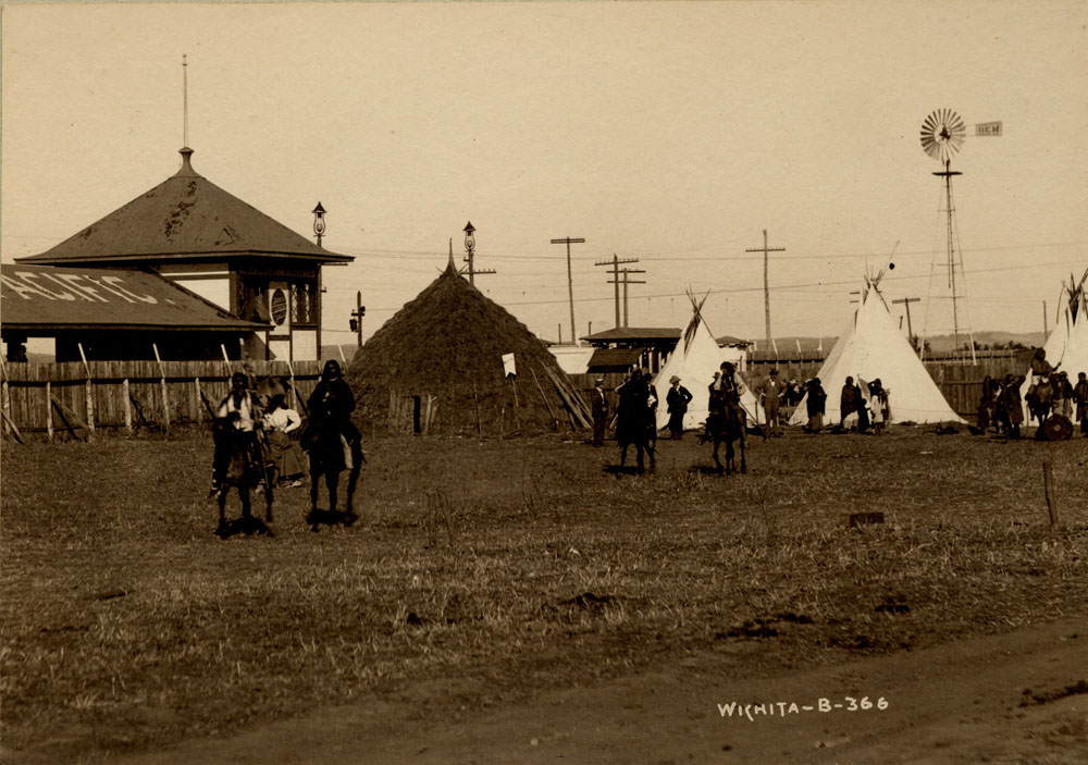 Wichita Delegates of the Indian Congress. Some on horseback near wigwam, others stand in groups by teepees with a few spectators. Missouri Pacific Railroad Station and windmill in background.