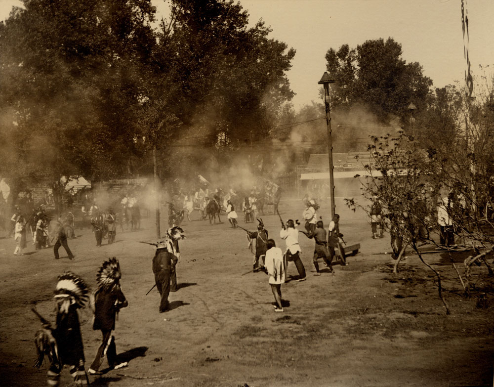 Indian Congress Participants engaged in mock battle. Gun smoke fills the air. Bleachers and building in the background.