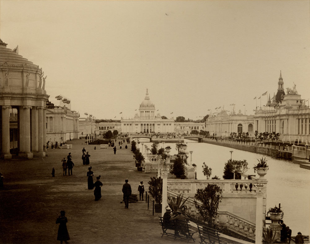 Grand Court looking west. Mines and Mining, Liberal, and Fine Arts Buildings to the north. Double bridge with balustraded island, U. S. Government Building and colonnades to the west. Agriculture, Administration arch, and Manufactures Buildings to the north. Balustrated staircases leading to Lagoon in front of Mines and Mining Building. Light crowd walking, seated, and standing.