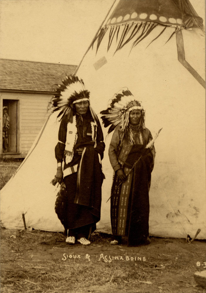 Group Portrait of Indian Congress Participants. Sioux and Assinaboine Delegates. Afraid of Eagle (Sioux) and Bear Cub (?) (Assiniboine) stand in front of teepee. An unidentified man stands in doorway of building in background.
