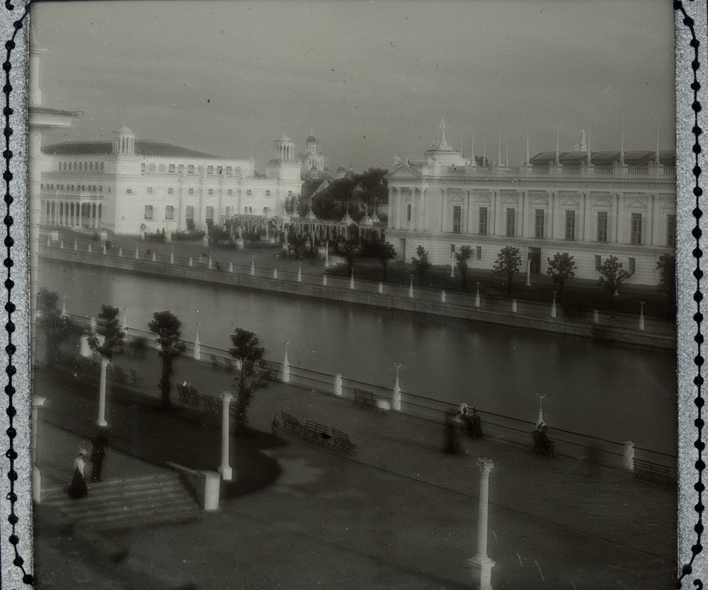 Mines and Mining Building and Liberal Arts Building with connecting colonnade and courtyard, looking southeast from across lagoon. A few people walking. Horticulture building visible in distance to the southeast. Stairs in front of Manufactures Building in foreground. Made from F. A. Rinehart photograph.