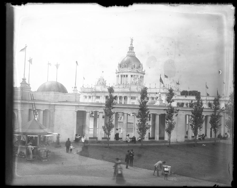 U.S. Government Building, looking southwest from behind West Colonnade. People lounging on stairs of colonnade. Produce stand on north end of colonnade next to step ladder.