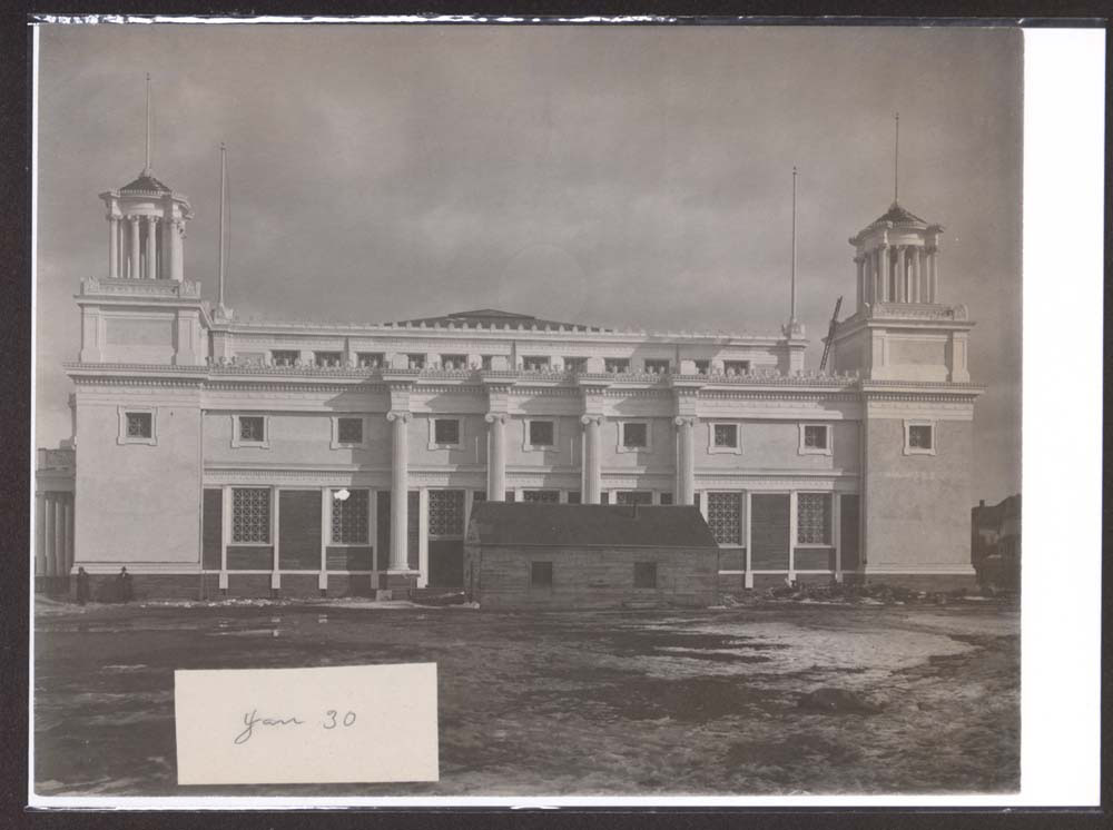 Construction of Mines and Mining Building. Two men stand at front left of structure. Snow on the ground.
