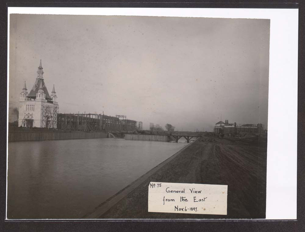 View of Administration Arch and Manufacturers Building Construction looking northeast from across lagoon. Mines and Mining Building Construction to the southeast. Construction of balustraded island. Water in lagoon.
