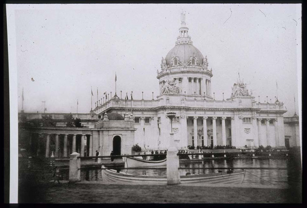 U.S. Government Building and portion of south west colonnade. Crowd seated on steps. Lagoon with docked boats.
