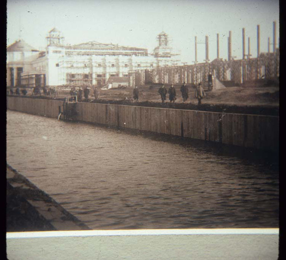Construction of Mines and Mining Building and Liberal Arts Building, looking southeast. Crowd walks along top of retaining wall lining lagoon.
