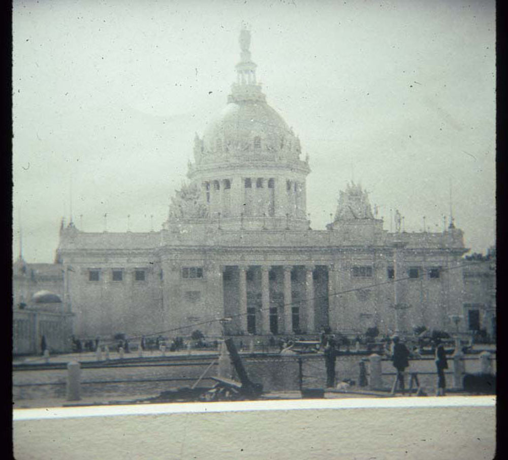Construction of U.S. Government Building. People stand along railing of lagoon. Swan boat and gondolas in water.